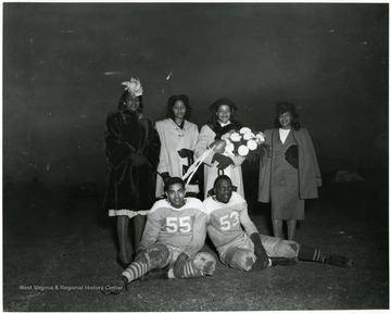 Four female students standing in the back row.  Two football players seated on the ground.